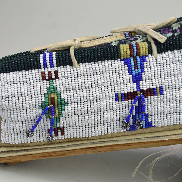 This Cheyenne-style toy cradle features wood base and a rawhide platform, fully beaded with 13/0 cut seed beads and secured with sinew stitching—showcasing traditional craftsmanship in miniature form. An up-close view of the beadwork reveals the intricate patterns and vibrant colors that reflect tribal identity and spiritual meaning