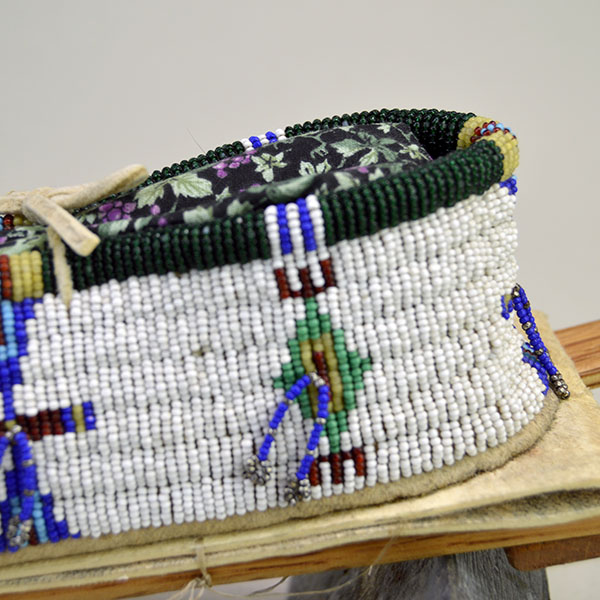 This Cheyenne-style toy cradle features wood base and a rawhide platform, fully beaded with 13/0 cut seed beads and secured with sinew stitching—showcasing traditional craftsmanship in miniature form. An up-close view of the beadwork reveals the intricate patterns and vibrant colors that reflect tribal identity and spiritual meaning