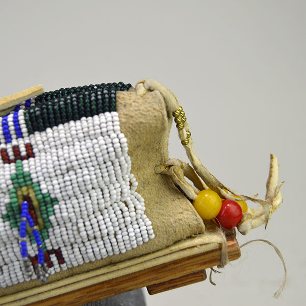This Cheyenne-style toy cradle features wood base and a rawhide platform, fully beaded with 13/0 cut seed beads and secured with sinew stitching—showcasing traditional craftsmanship in miniature form. An up-close view of the back beadwork reveals the intricate patterns and vibrant colors that reflect tribal identity and spiritual meaning.