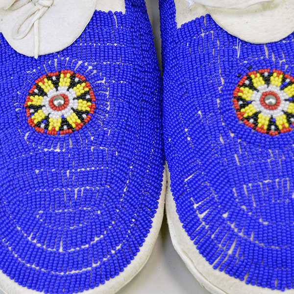 The striking blue beaded moccasins, crafted from soft white deerskin and sewn with thread, feature durable white strap leather soles and are fully adorned in a lazy stitch beadwork pattern, beautifully showcased in close-up pictures that highlight the intricate detail.