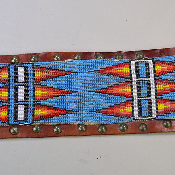 This vintage handcrafted belt, beaded in blue with a geometric design, is pictured here showcasing its intricate beadwork along with the .25-inch brass tacks and .5-inch brass spots