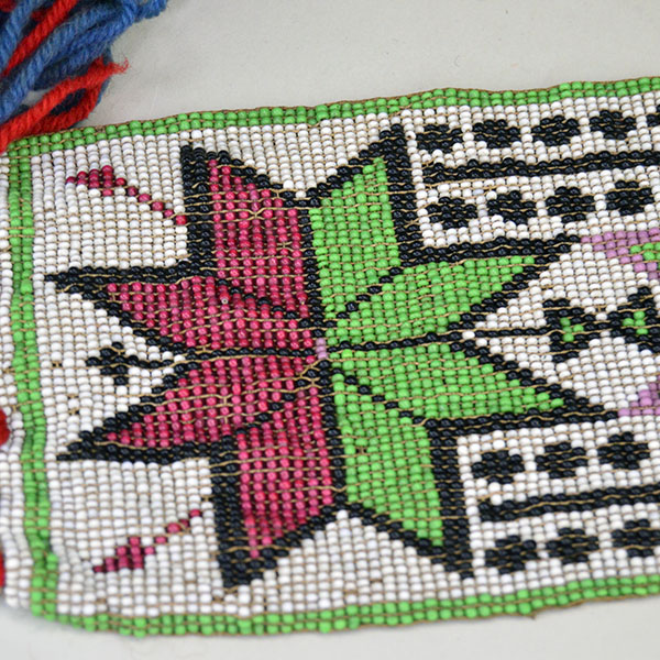 Sash- Antique Loom Beaded showcases a bold floral design. Approx. 24" partially braided wool‑yarn fringe. the sash reaches approx. 55" in total length. a red and green beaded flower.