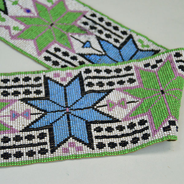 Sash- Antique Loom Beaded showcases a bold floral design. Approx. 24" partially braided wool‑yarn fringe. the sash reaches approx. 55" in total length. One of the blue beaded flowers.