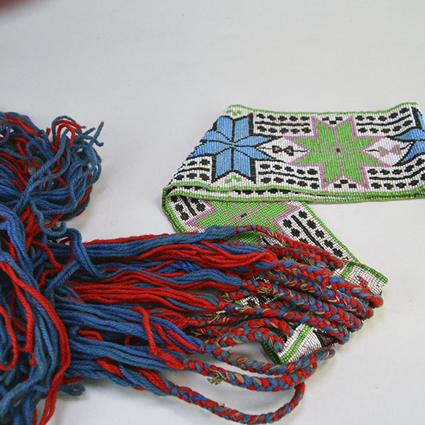 Sash- Antique Loom Beaded showcases a bold floral design. Approx. 24" partially braided wool‑yarn fringe. the sash reaches approx. 55" in total length. the blue and red fringe against the colorful bead work.