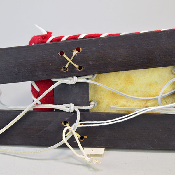 Cradleboard Parfleche features rawhide construction and a hand‑painted design. measures approximately 21 inches long, 7 inches wide at the head. How the skis and the cradleboard are attached with raw hide and deerskin leather lacing.
