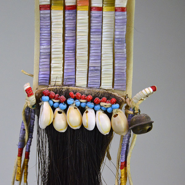 Wapegnaki- Hair Ornament Quilled features 9″ of quillwork on a 10″ rawhide base. Eight 5″ quilled tassels hang from the piece, 23″ dark horse‑hair drop. The pony beads and the cowrie shells before the hore hair drop.