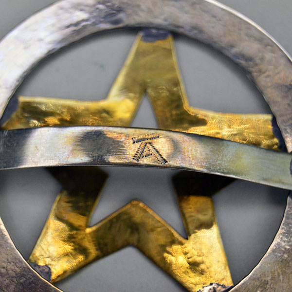 Hand‑stamped Brass Star Scarf Slide with German silver circle base. Measures 2.5″ x 2.5″. Bold, compact accessory for regalia, bandanas, and neckerchiefs. the A with the line over the top on the metal loop on the back.